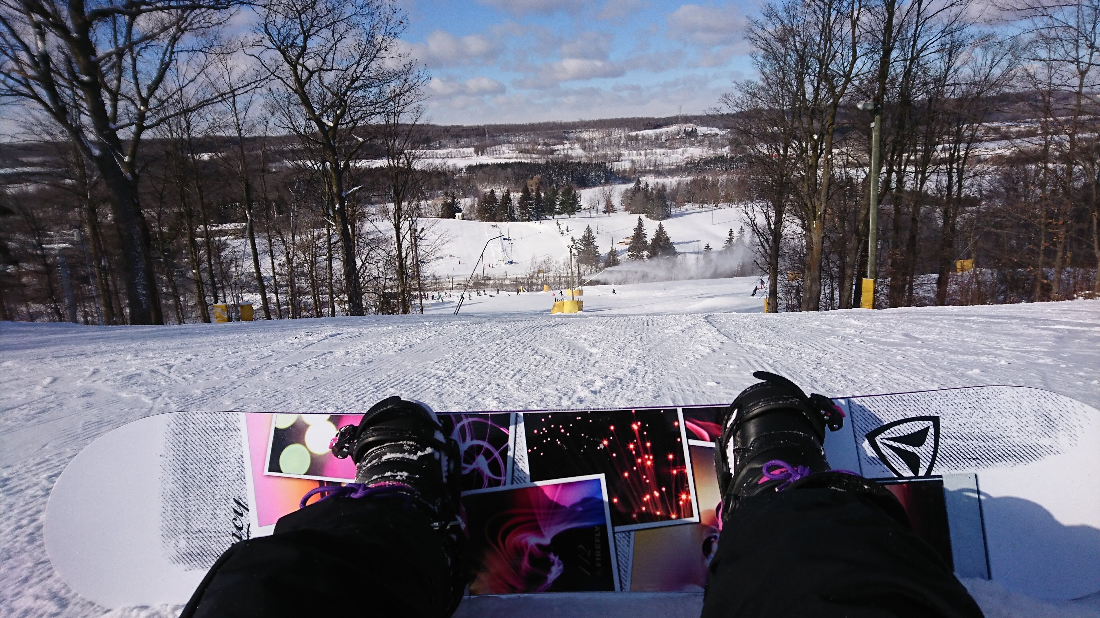 A snowboarder's feet on a snowboard with a view of a snowy landscape and a ski lift in the background.