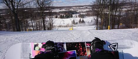 Les pieds d’un snowboarder sur une planche de snowboard, avec en arrière-plan un paysage enneigé et un télésiège.