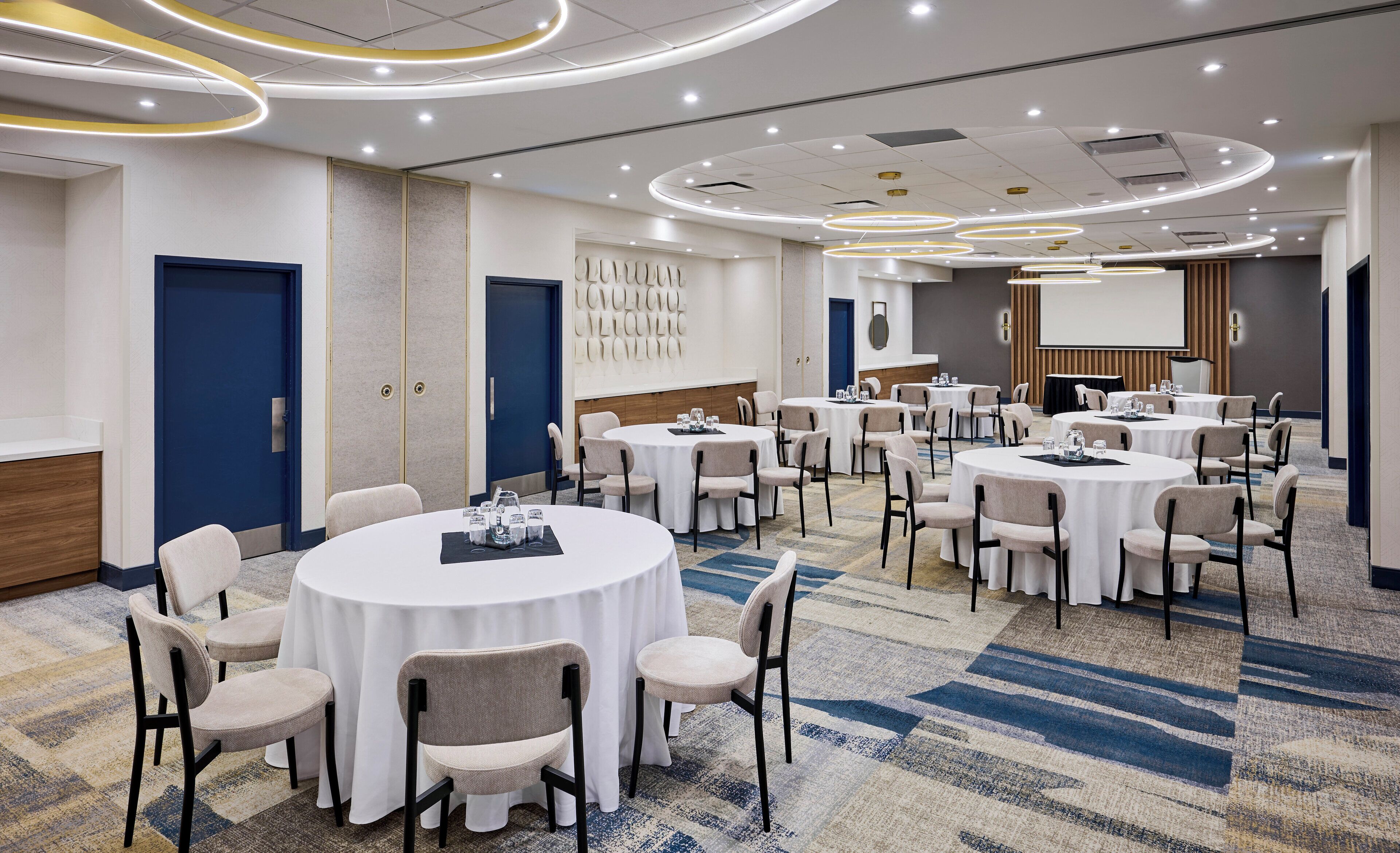 A conference room with multiple round tables, each with chairs, equipped with glassware and a projector screen.