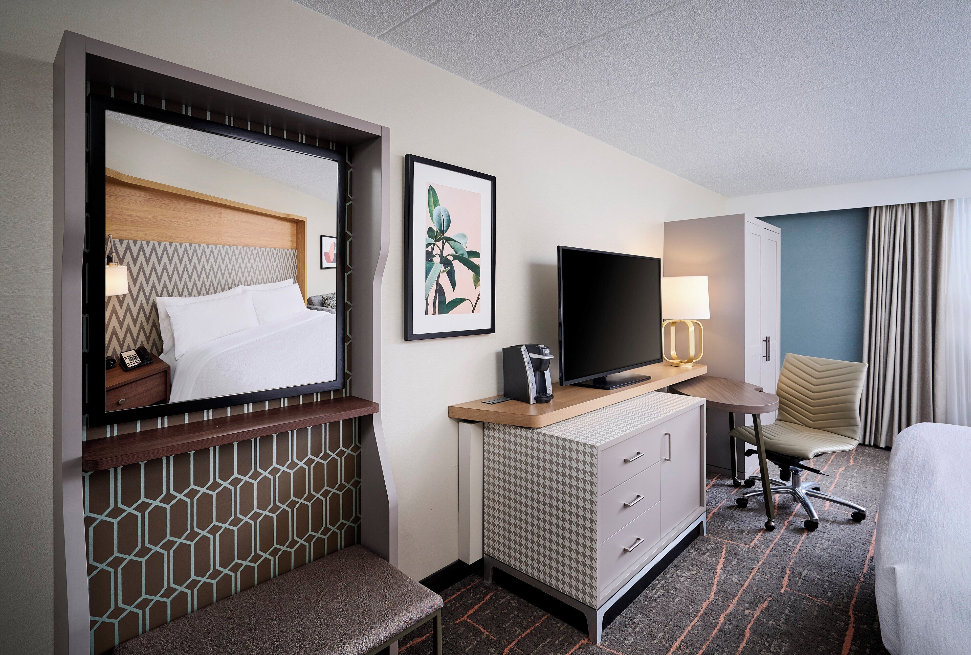A hotel room with a bed, a TV, a desk with a lamp, a chair, and a small table.