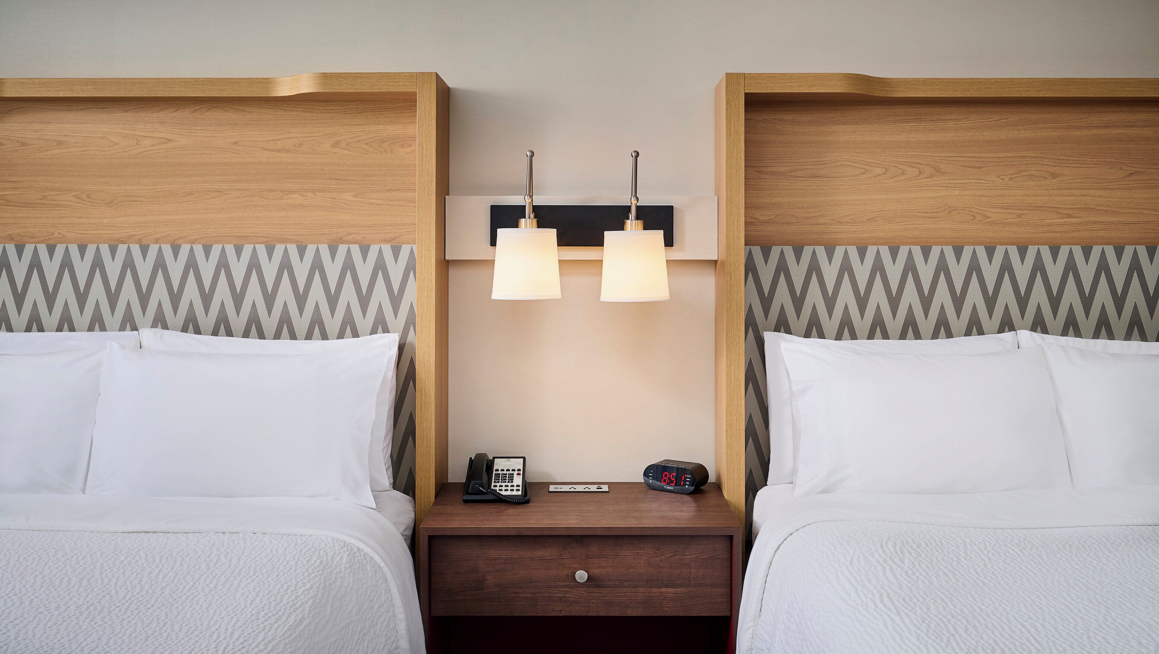 Habitación de hotel con dos camas, una mesita de noche con teléfono y reloj, y una lámpara de pared.