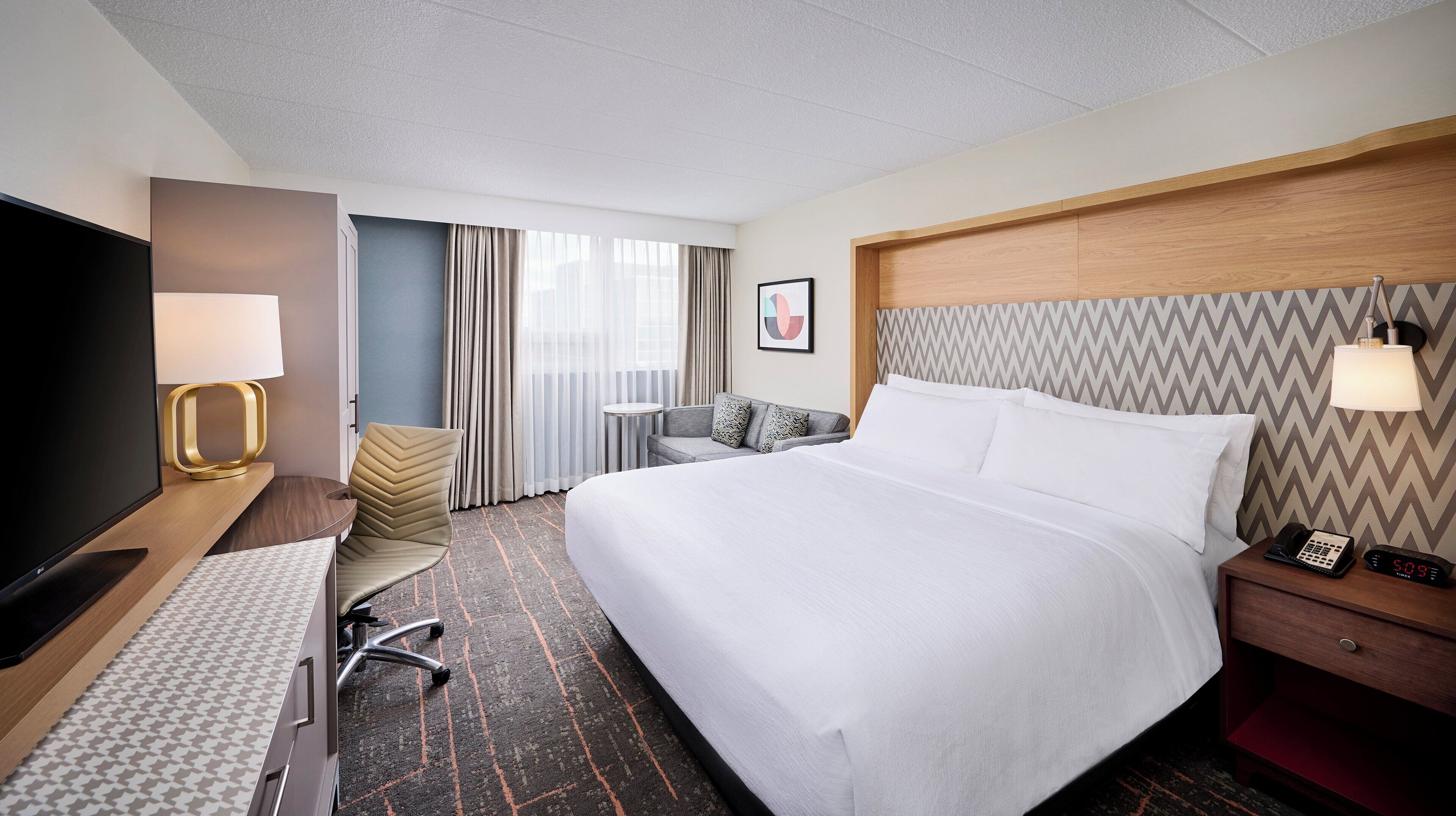 Una habitación de hotel con una cama grande, un escritorio con lámpara, televisión y una mesita de noche con teléfono y reloj.