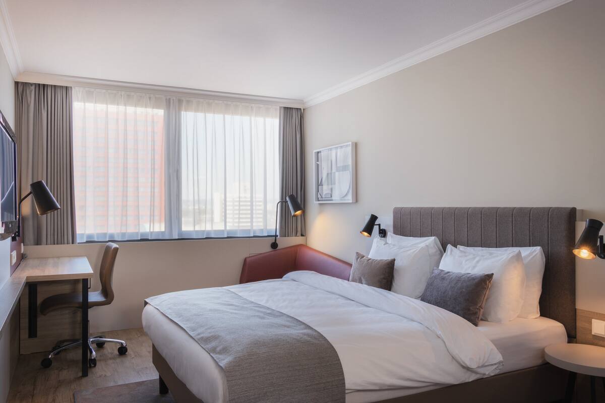 A hotel room with a bed, desk, chair, and a window with curtains.