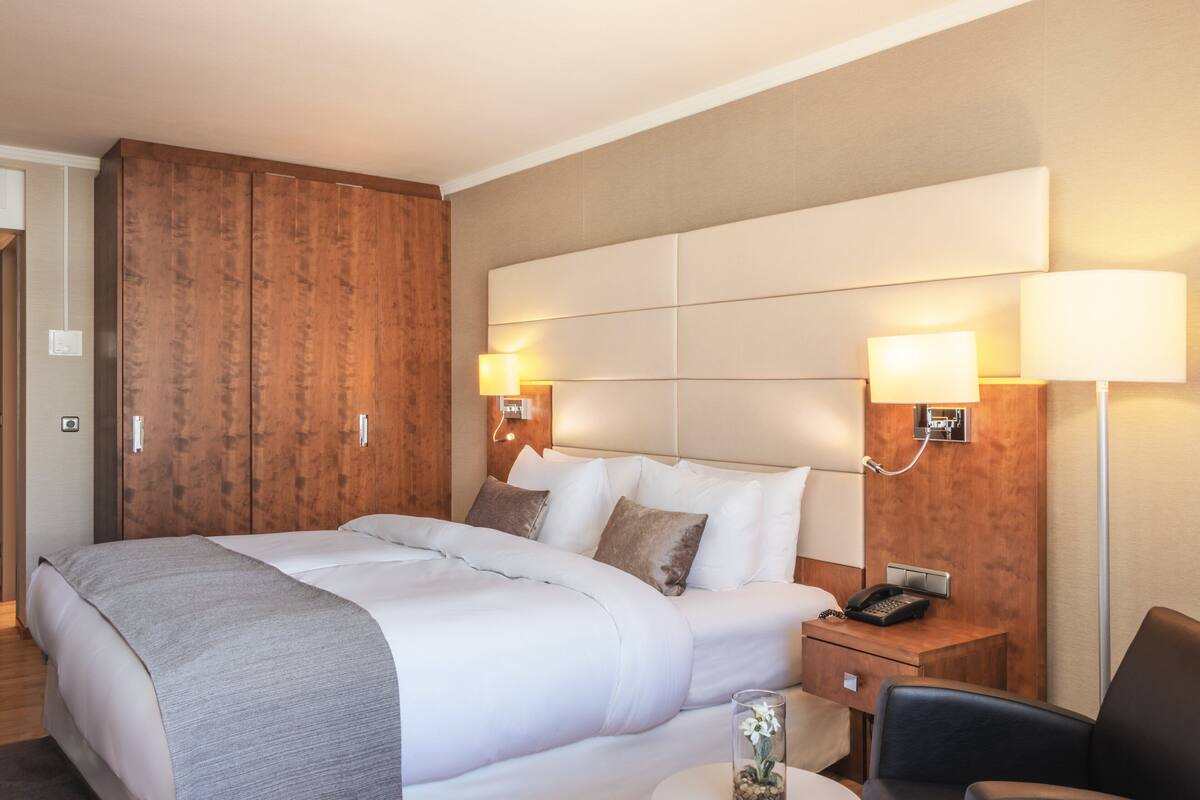 A hotel room with a large bed, wooden wardrobe, bedside lamps, a telephone, and a small table with a vase.