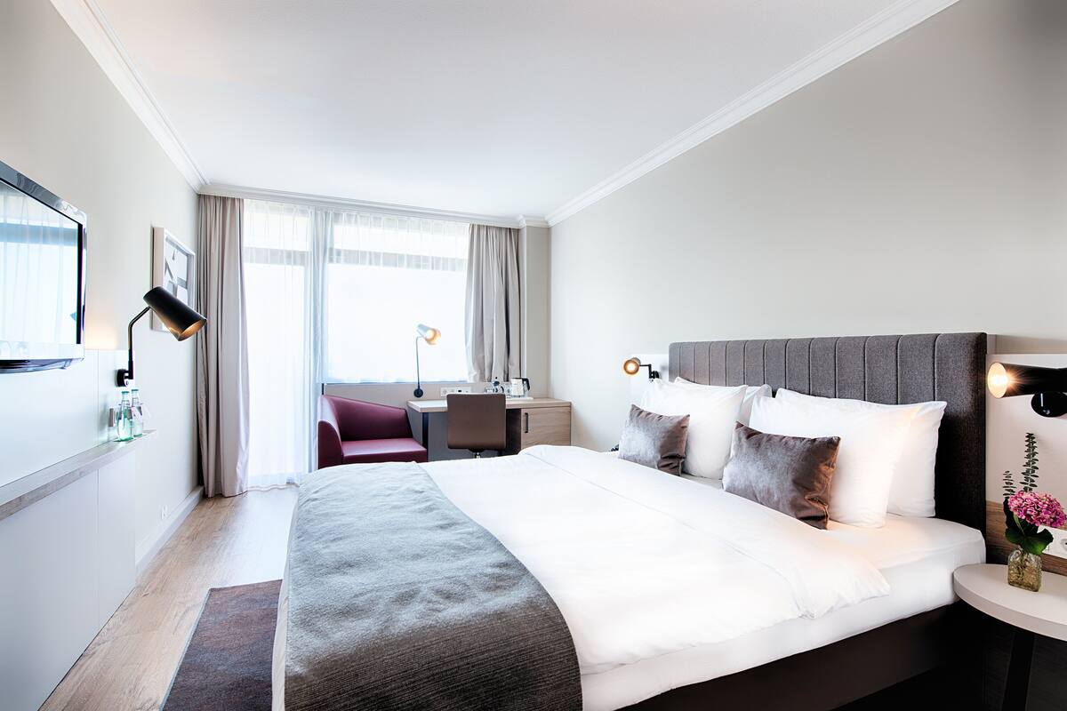 A hotel room with a large bed, a desk, a chair, and a TV on the wall.