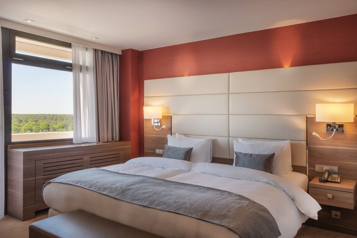 A hotel room with a large bed, two bedside tables, a nightstand, a window with curtains, and a view of greenery outside.