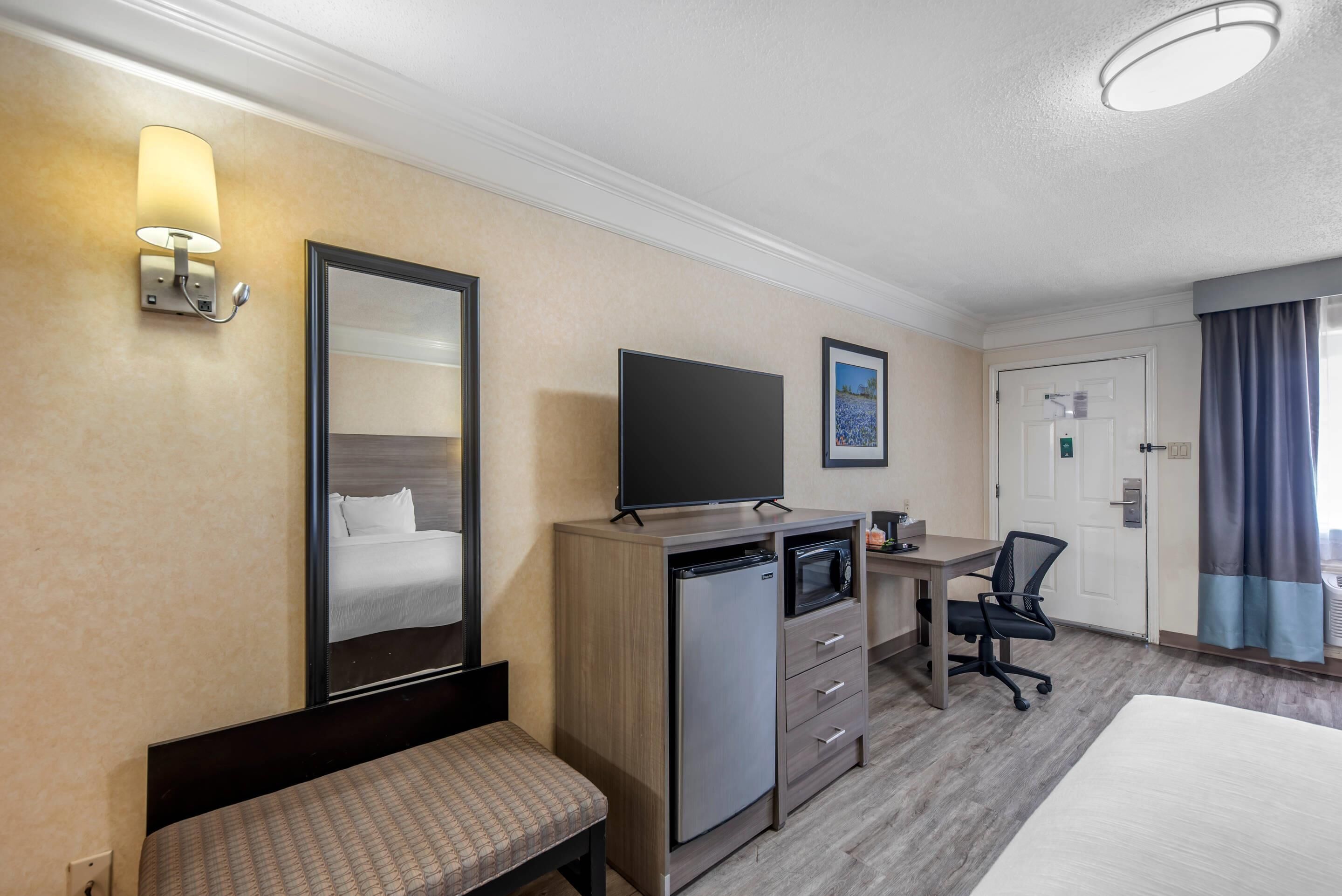 A hotel room with a bed, a desk with a computer, a refrigerator, a microwave, a chair, and a television.