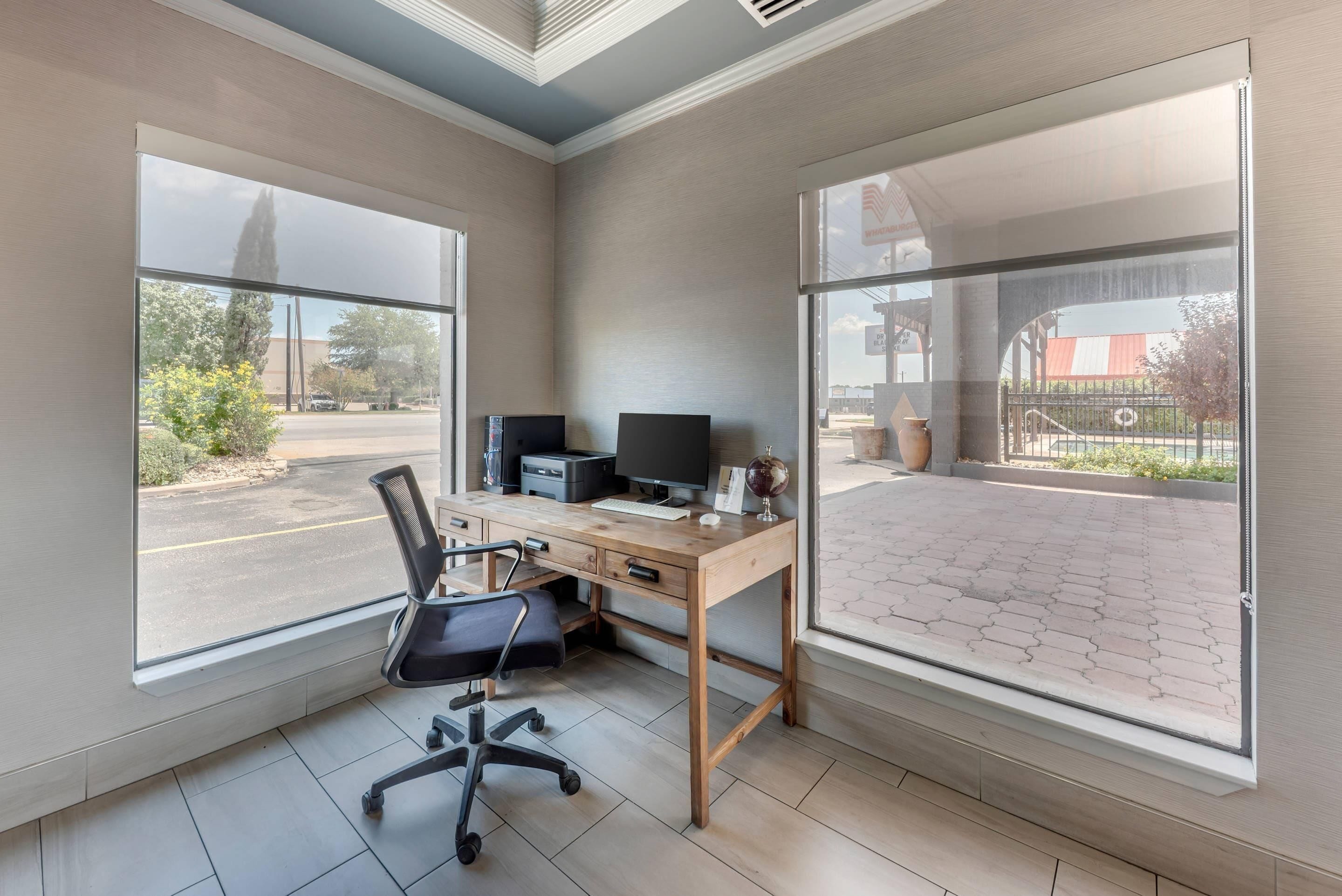 A modern office space with a wooden desk, black office chair, and large windows offering views of a parking lot and outdoor area.