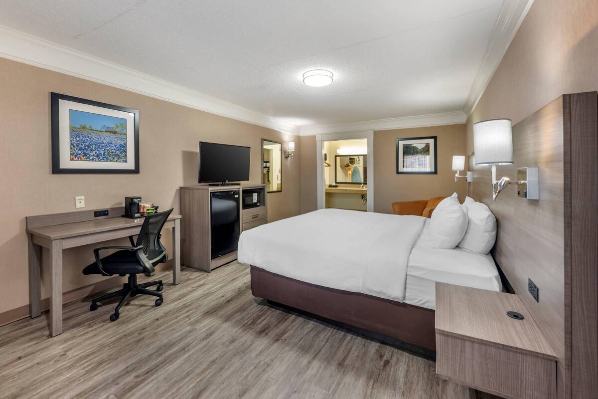 Une chambre d’hôtel avec un grand lit, un bureau avec un ordinateur, une télévision et une salle de bain visible à travers une porte ouverte.