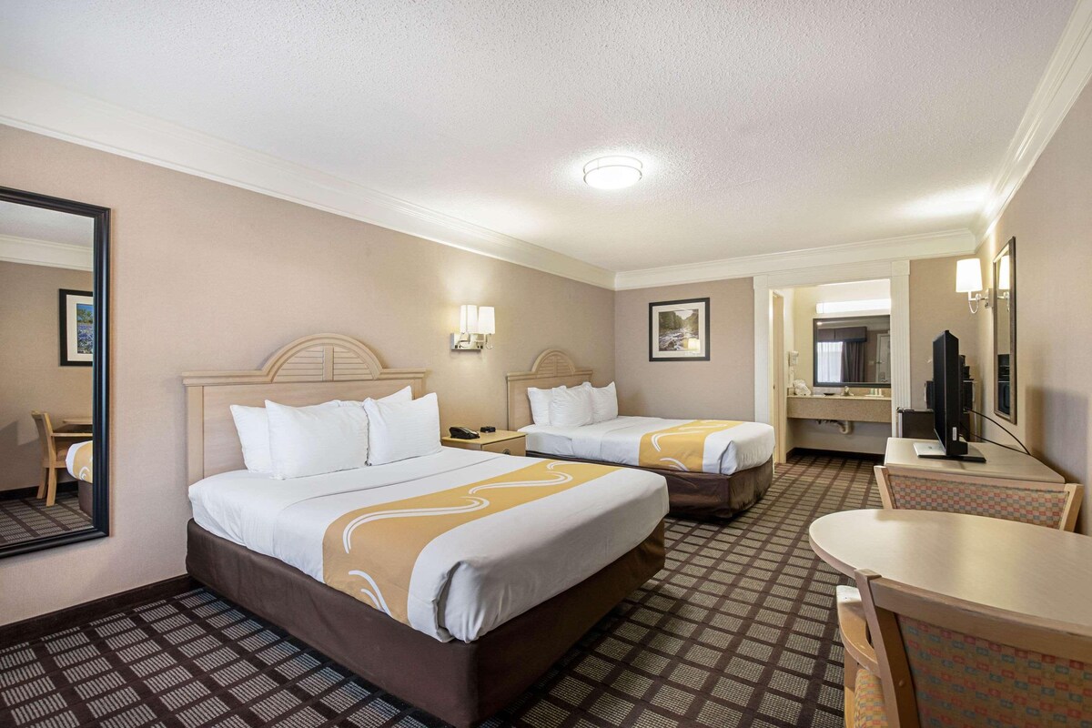 A hotel room with two beds, a desk, a chair, a TV, and a bathroom visible through an open door.