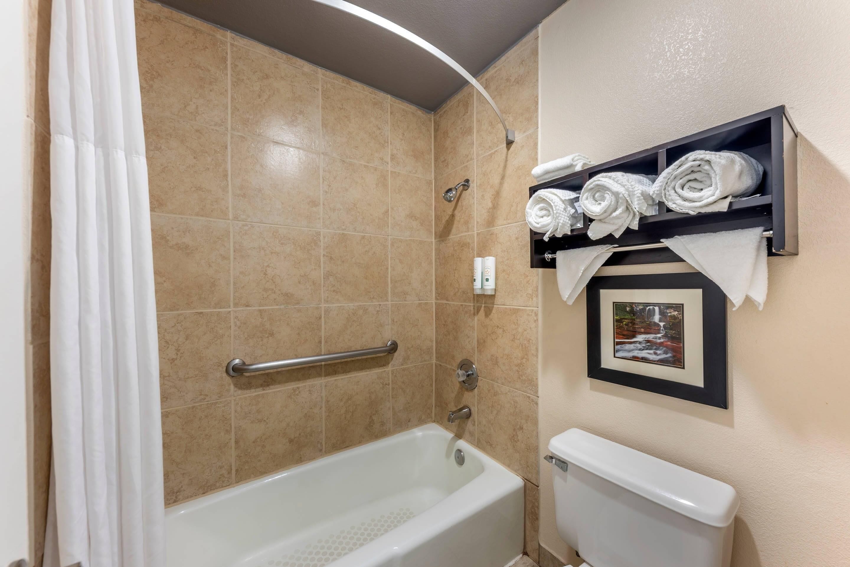 Un baño con bañera, inodoro, ducha con barra de apoyo y estante con toallas.