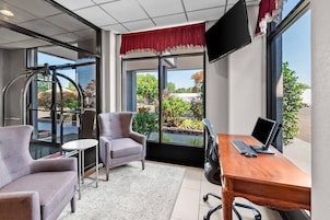 Habitación de hotel con escritorio, dos sillas, televisor y ventanas grandes con vistas a zonas verdes.
