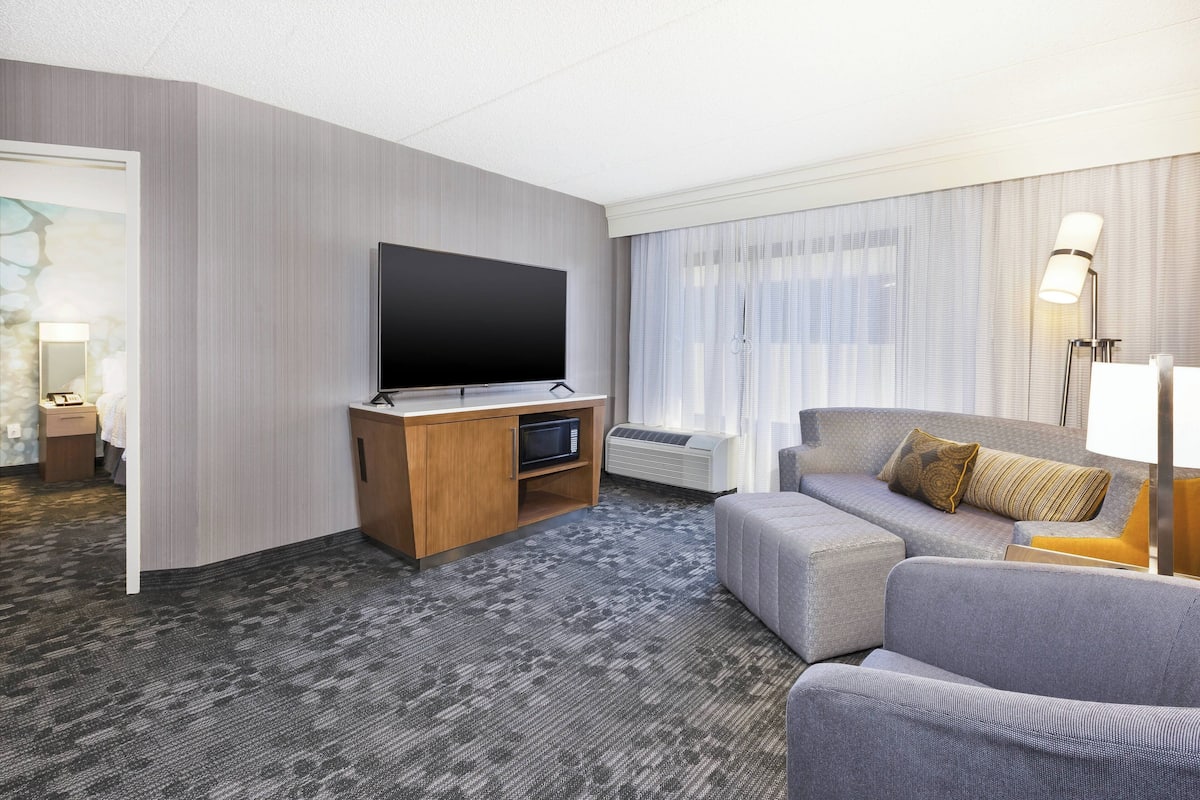 Een moderne hotelkamer met een flatscreen-tv, een bank, een bureau met een magnetron, en een groot raam met gordijnen.