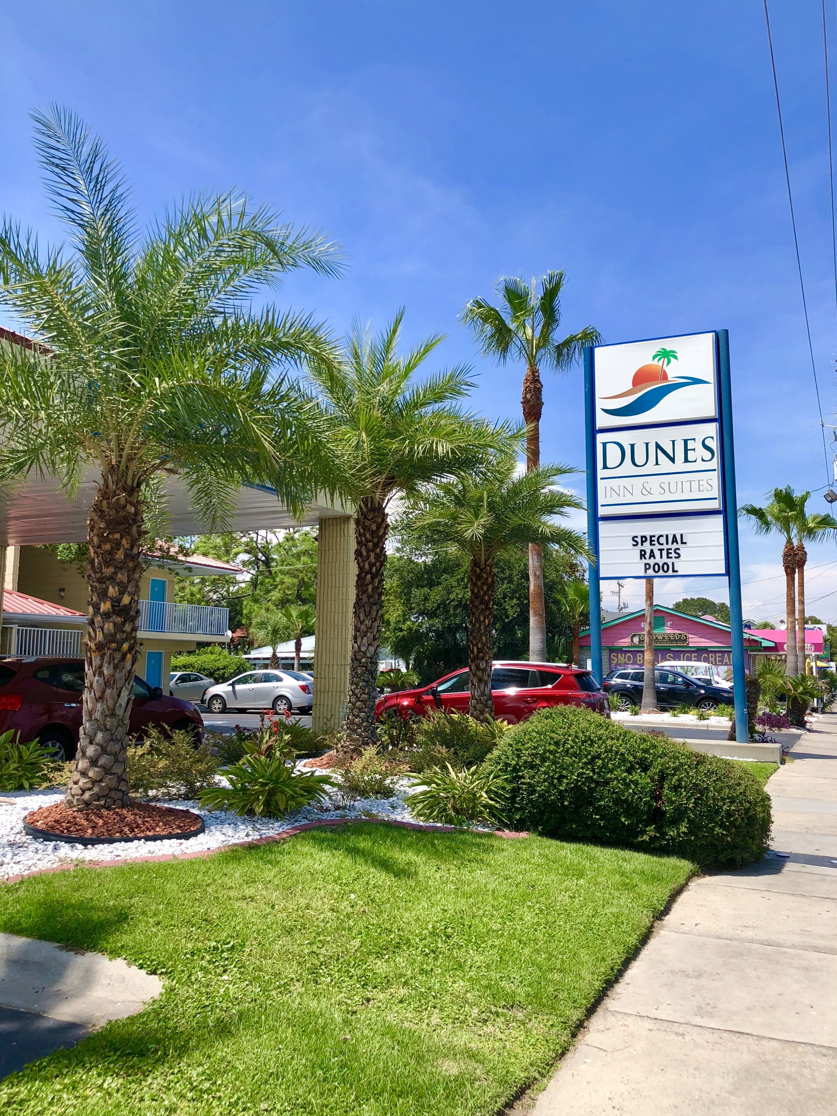 A Dunes Inn & Suites sign with a palm tree and beach logo, surrounded by palm trees and a parking lot with cars.