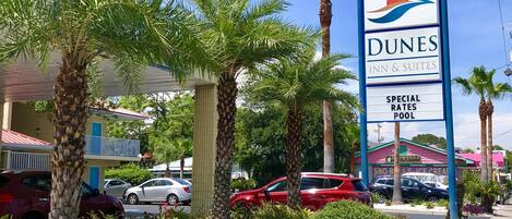 A Dunes Inn & Suites sign with a palm tree and beach logo, surrounded by palm trees and a parking lot with cars.