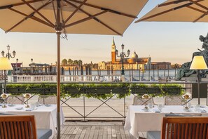 An outdoor dining area with tables set for two, overlooking a waterfront with a historic building in the background. - Londra Palace Venezia (Venice)