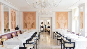 Uma sala de jantar formal com mesas longas, cadeiras elegantes e um lustre imponente.