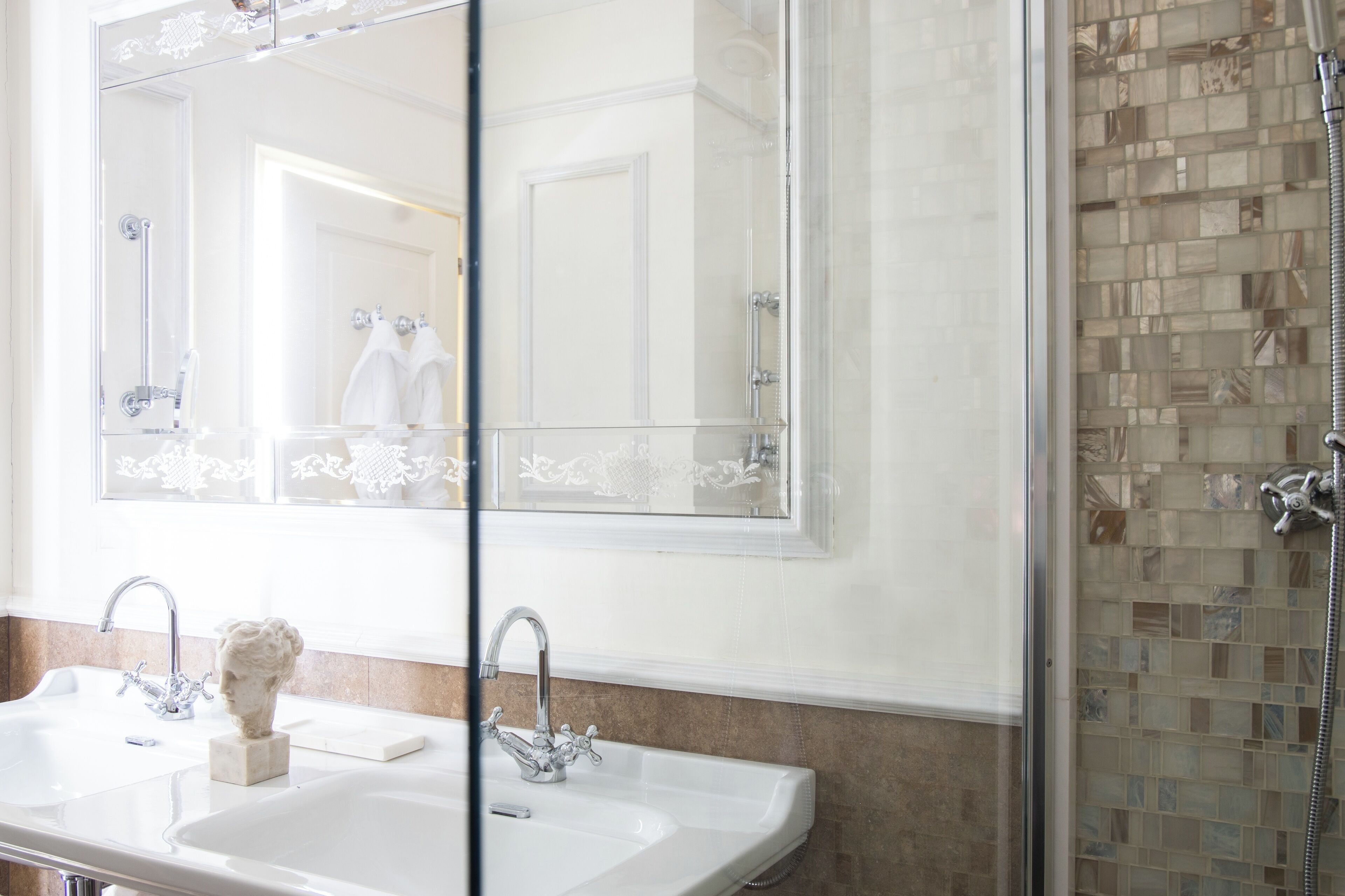 Un baño con un espejo grande, dos lavamanos, un busto de mármol y una ducha con mosaicos.