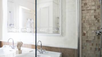 Un baño con un espejo grande, dos lavamanos, un busto de mármol y una ducha con mosaicos.
