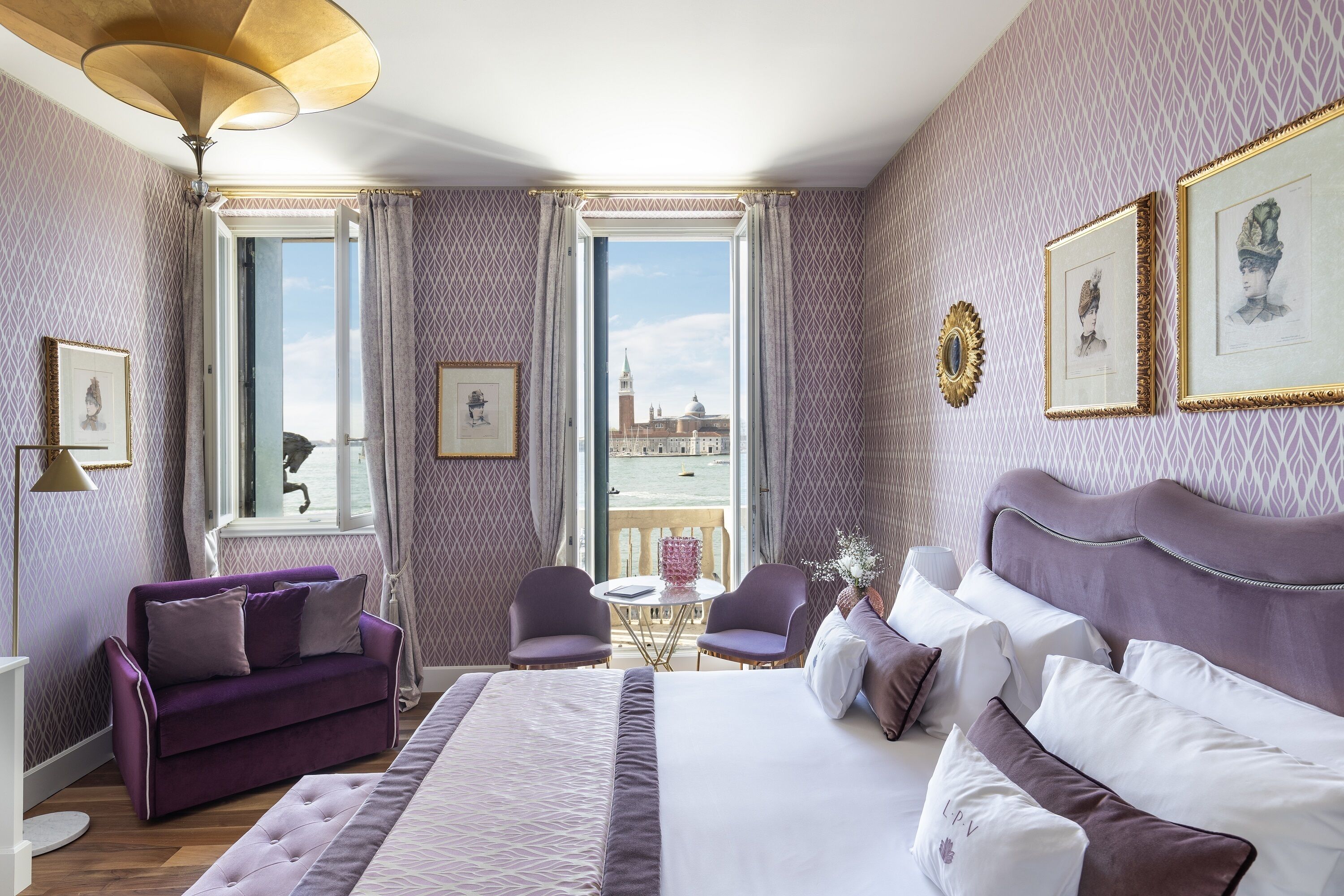 A hotel room with a large bed, a purple sofa, two armchairs, a small round table, and a view of the cityscape through the windows.