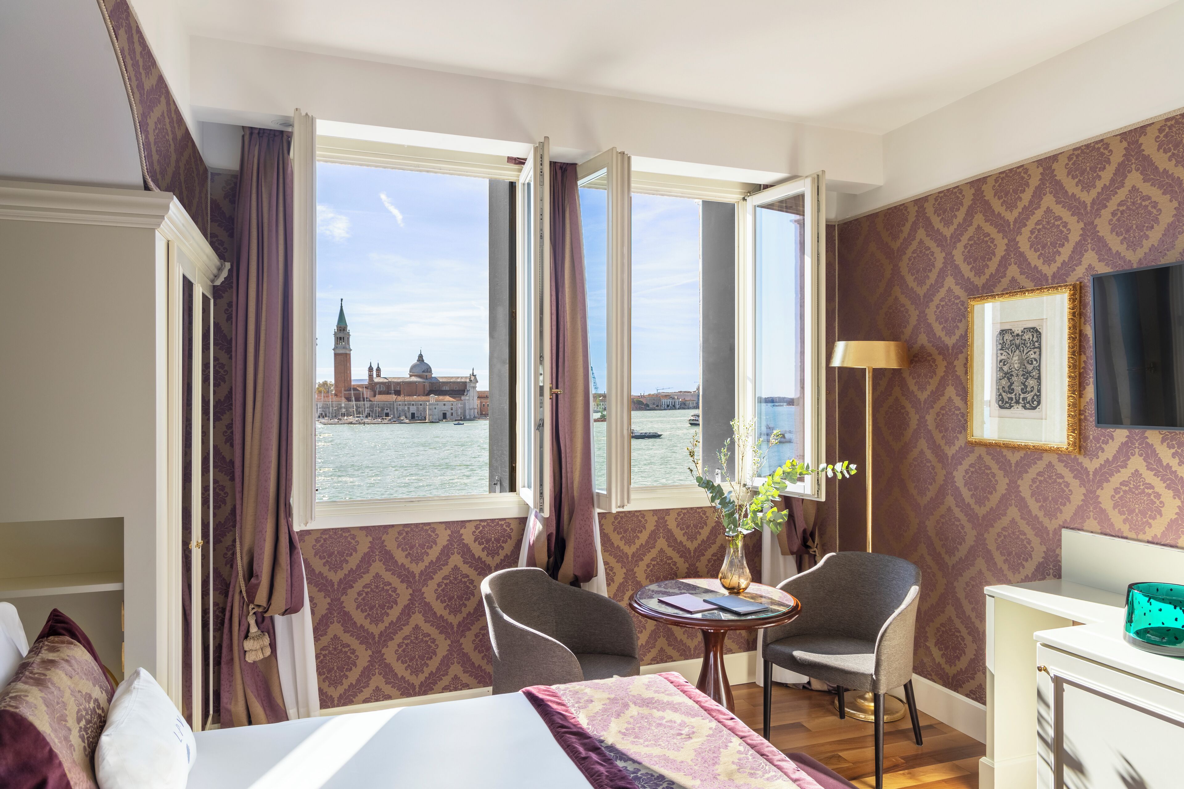 A hotel room with a bed, two chairs, a table, and a desk. A large window offers a view of a historic building and a body of water.