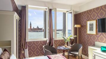 A hotel room with a bed, two chairs, a table, and a desk. A large window offers a view of a historic building and a body of water.