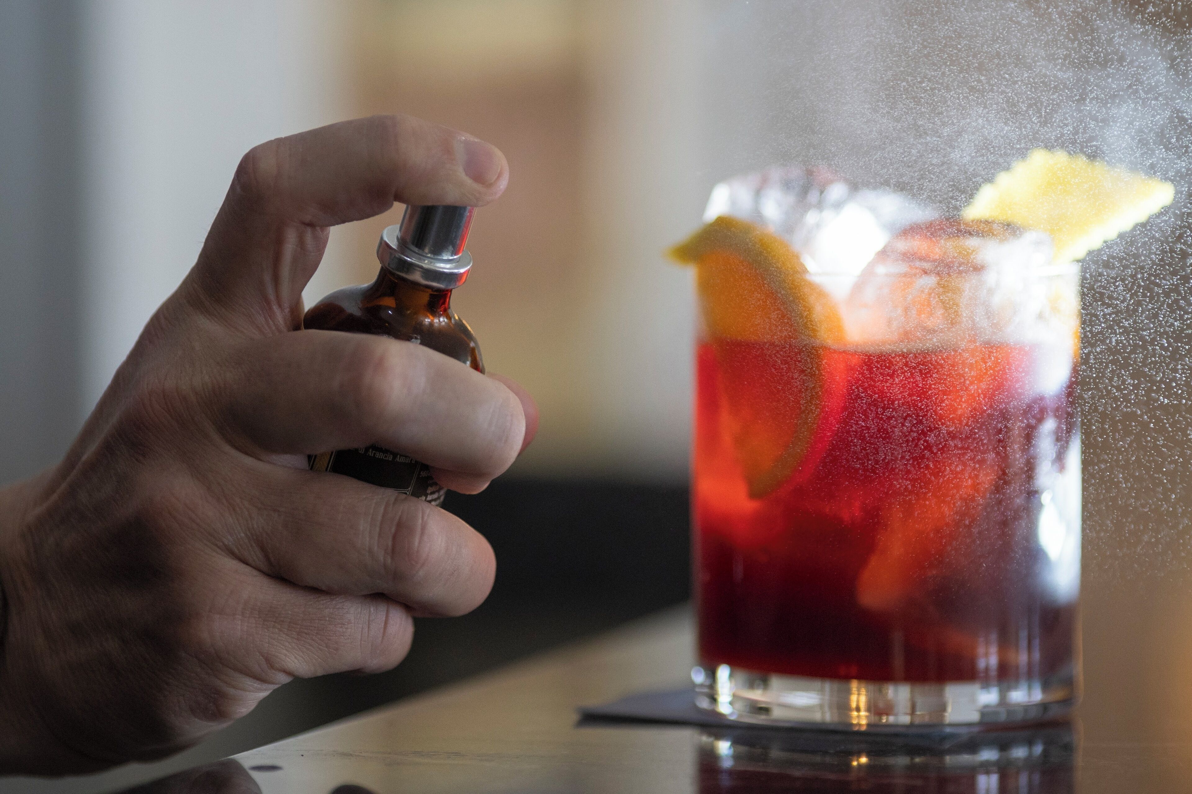 A hand holding a bottle, possibly a drink, near a glass with a red liquid and garnishes.