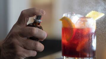 A hand holding a bottle, possibly a drink, near a glass with a red liquid and garnishes.
