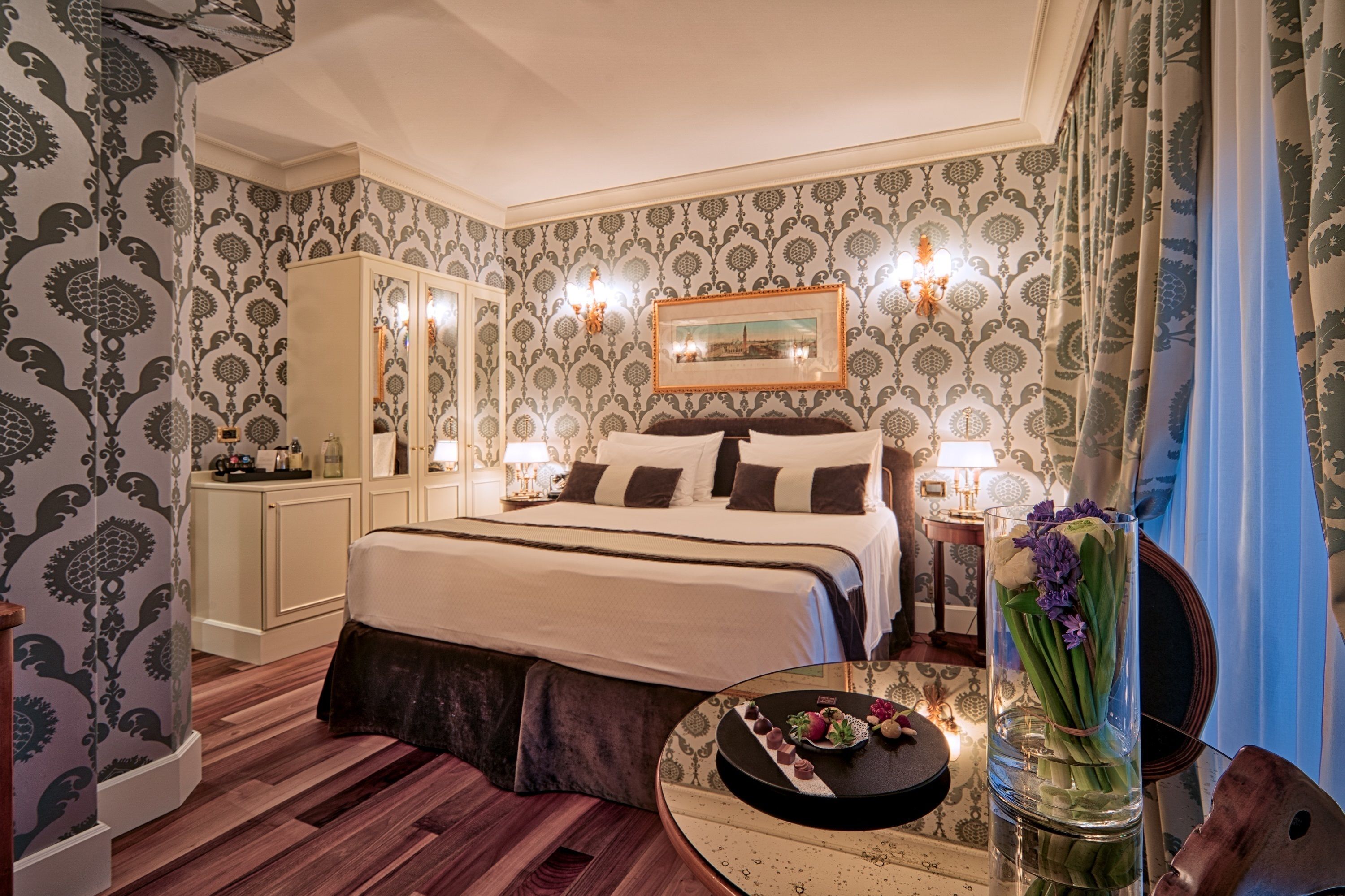 A bedroom with a large bed, a bedside table with a vase of flowers, and patterned wallpaper.