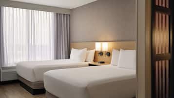 Habitación de hotel con dos camas, un ventanal con cortinas, una lámpara fijada a la pared y una puerta con persianas verticales.