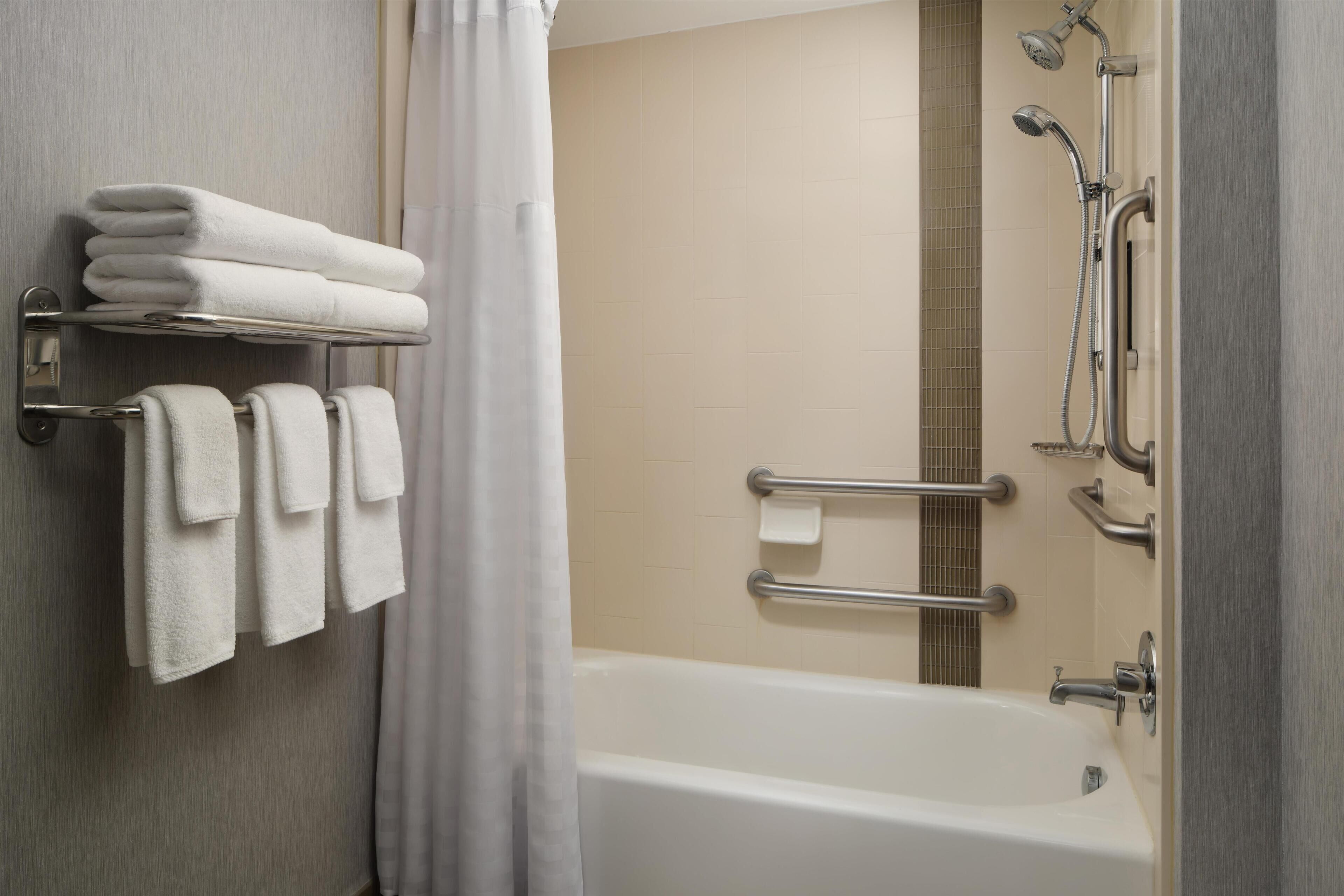 Un baño con bañera, un perchero con toallas dobladas, ducha con cabezal de ducha manual y barra de apoyo.