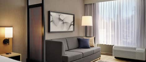 Una habitación de hotel moderna con un sofá gris, una alfombra estampada, un amplio ventanal con cortinas, una obra de arte en la pared y una lámpara de noche.