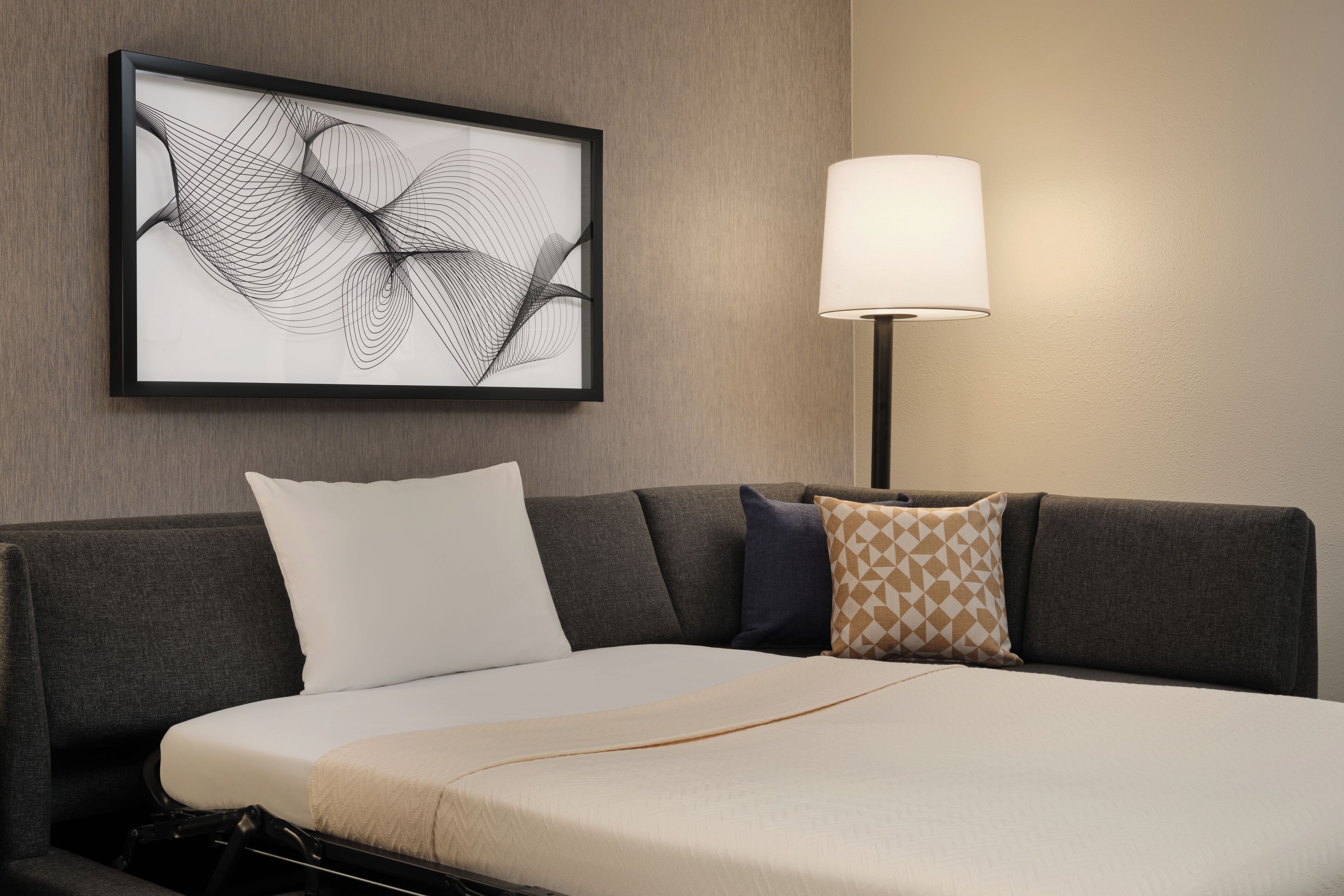 Una habitación de hotel moderna con una cama grande, una obra de arte abstracta enmarcada, una lámpara de pie y un sofá con cojines estampados.