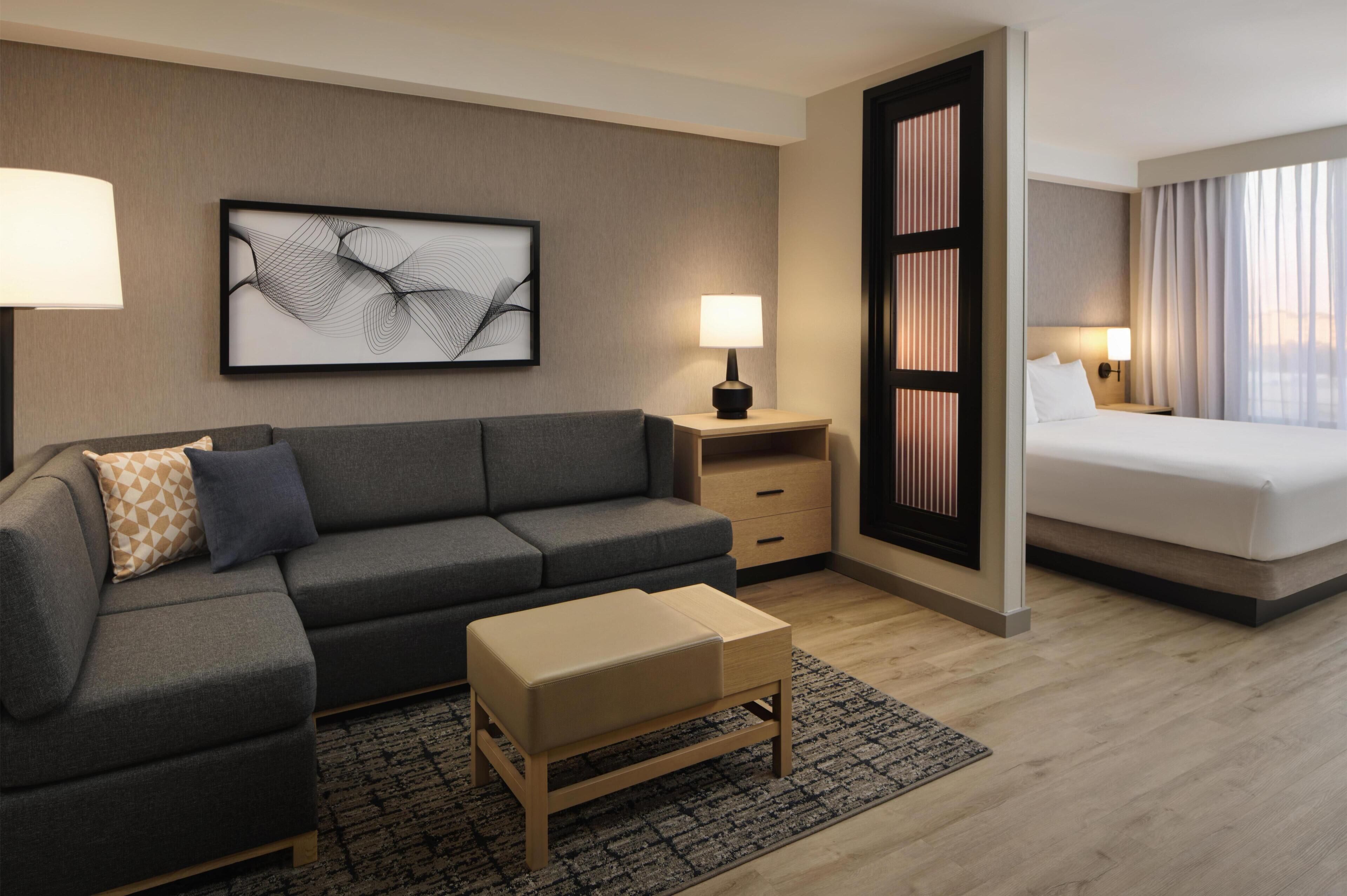 Habitación de hotel moderna con sofá gris, una pequeña mesa de madera, cama con ropa blanca, mesita de noche con lámpara y una obra de arte abstracto enmarcada en la pared.