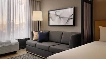 A hotel room with a gray sofa, a bed, a framed abstract artwork, a standing lamp, and a window with sheer curtains.