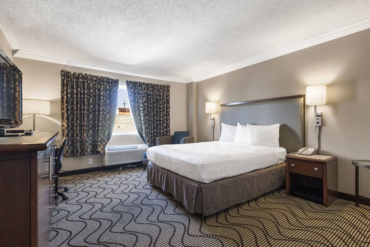 A hotel room with a large bed, a desk with a lamp, a chair, a TV, and a window with curtains.