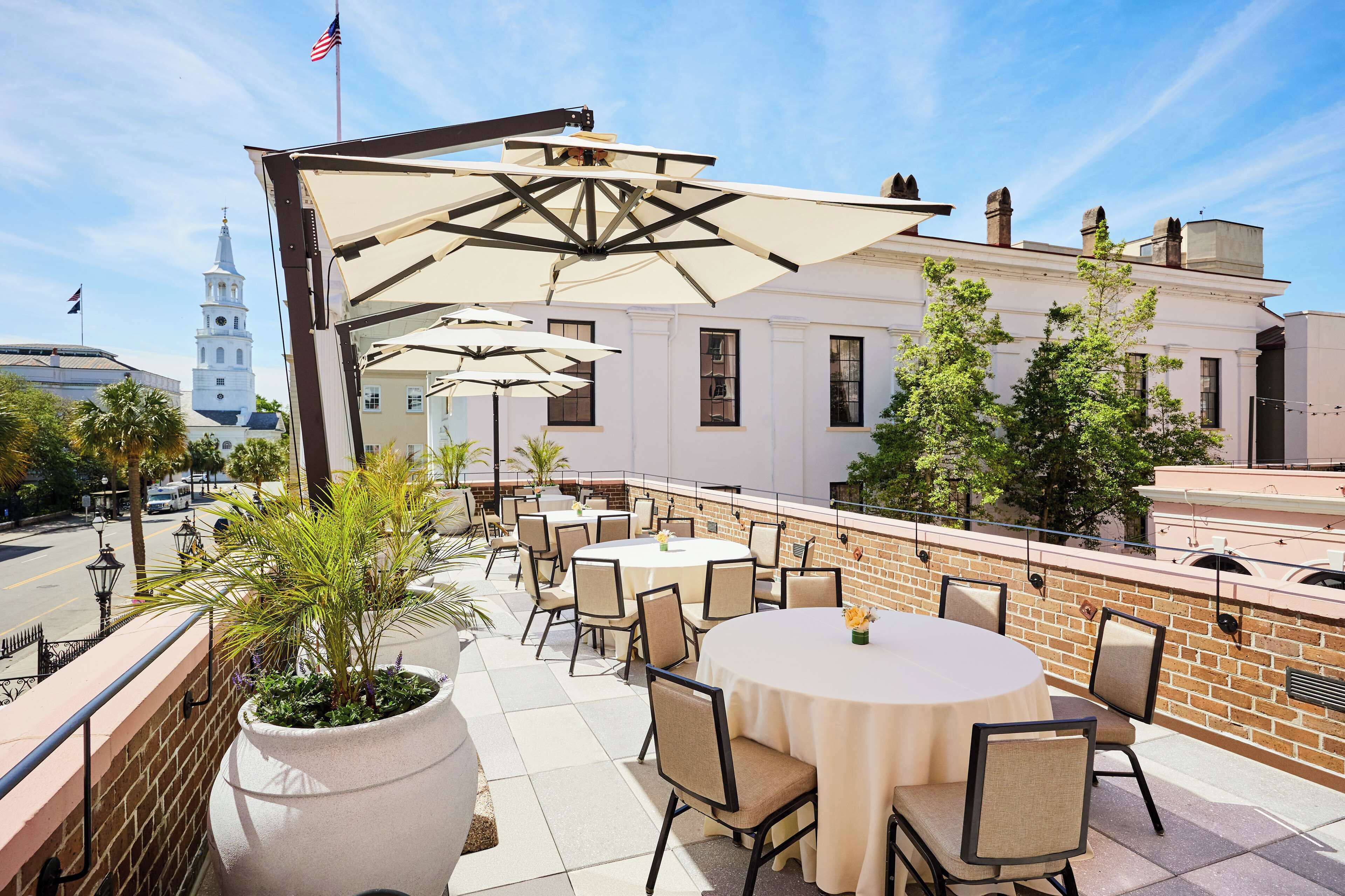 An outdoor patio with round tables and chairs, a brick wall, and a view of a white building with a clock tower.