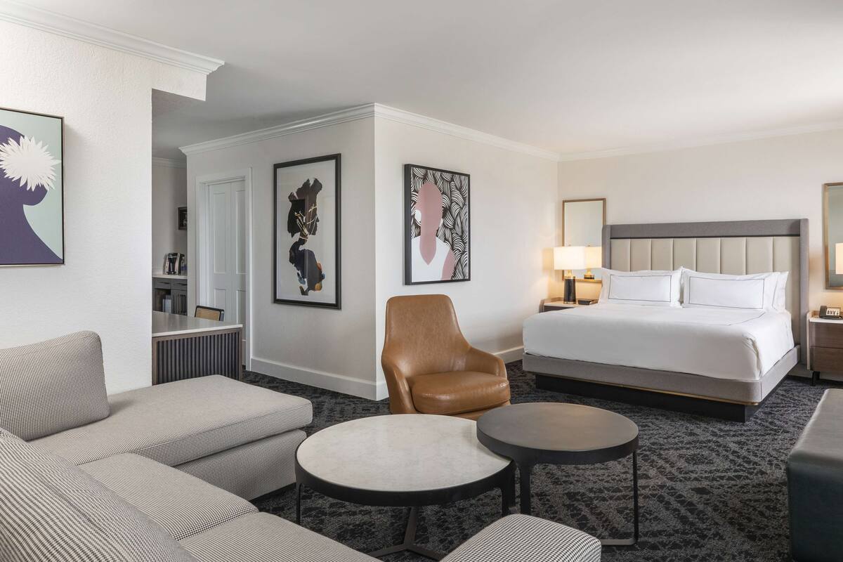 A hotel room with a bed, a sofa, a chair, a small table, and artwork on the walls.
