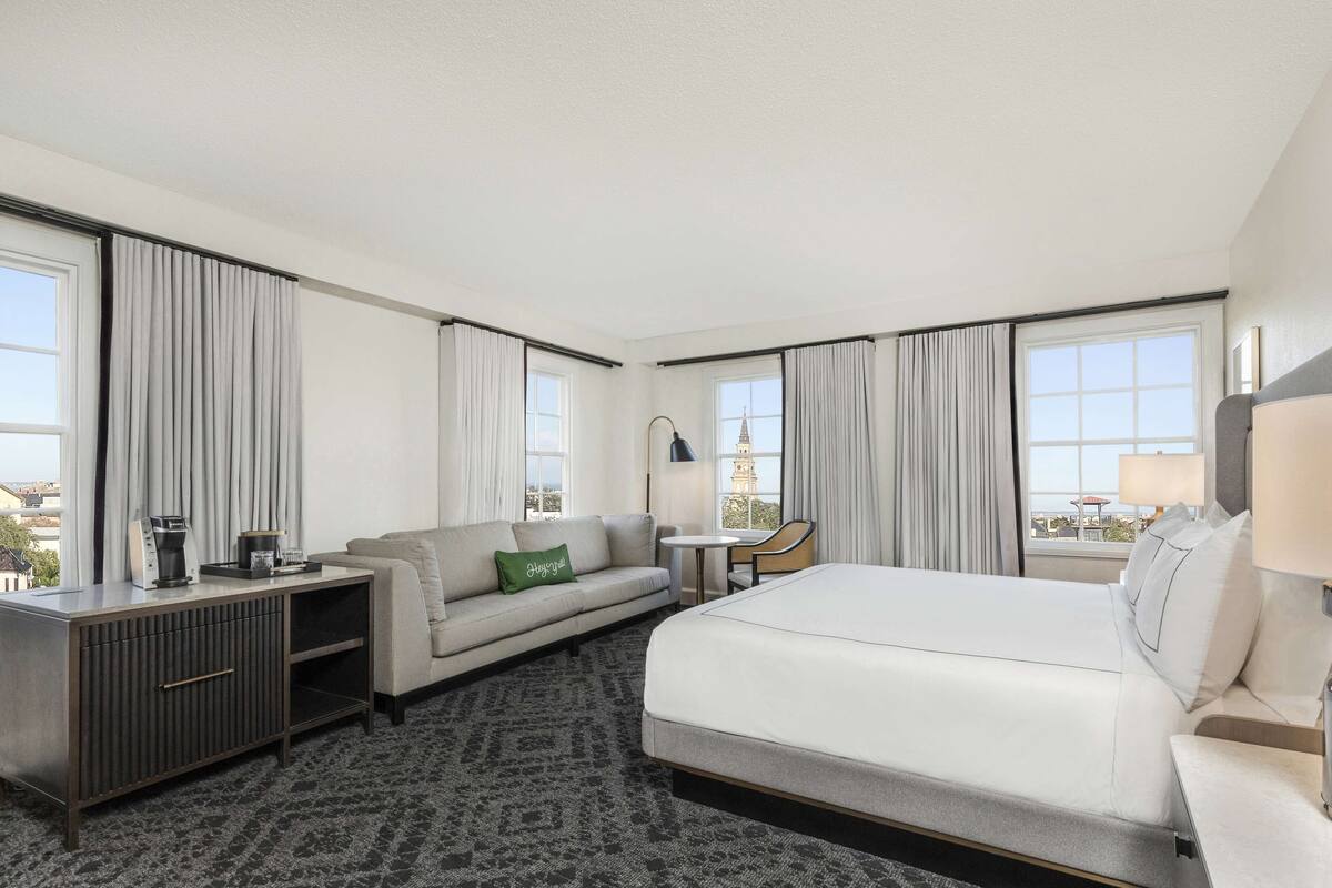 A hotel room with a bed, a sofa, a desk, and a chair. There are curtains on the windows, and a lamp on the desk.