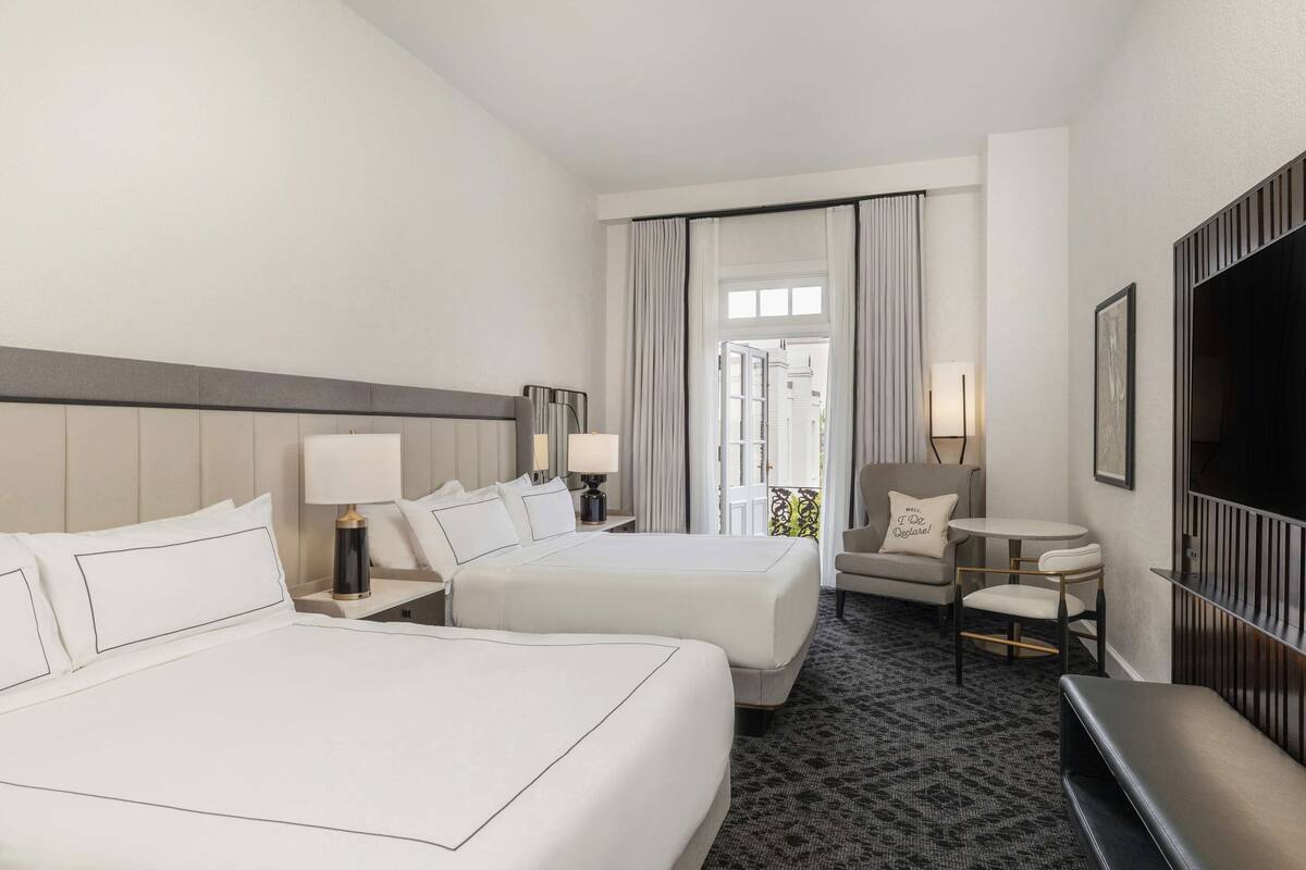 A hotel room with two beds, a small table, a chair, a TV, and a balcony with curtains.