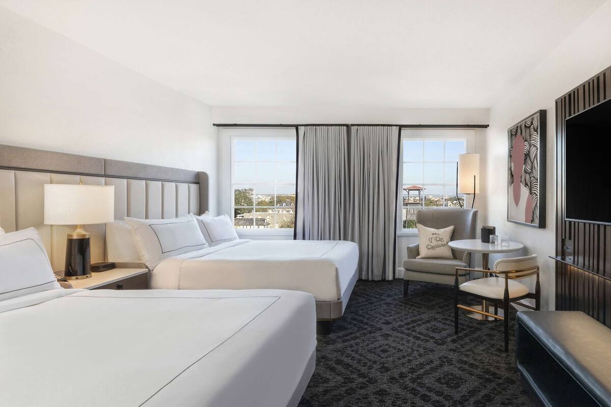 A hotel room with two beds, a desk, a chair, a TV, and a large window with curtains.