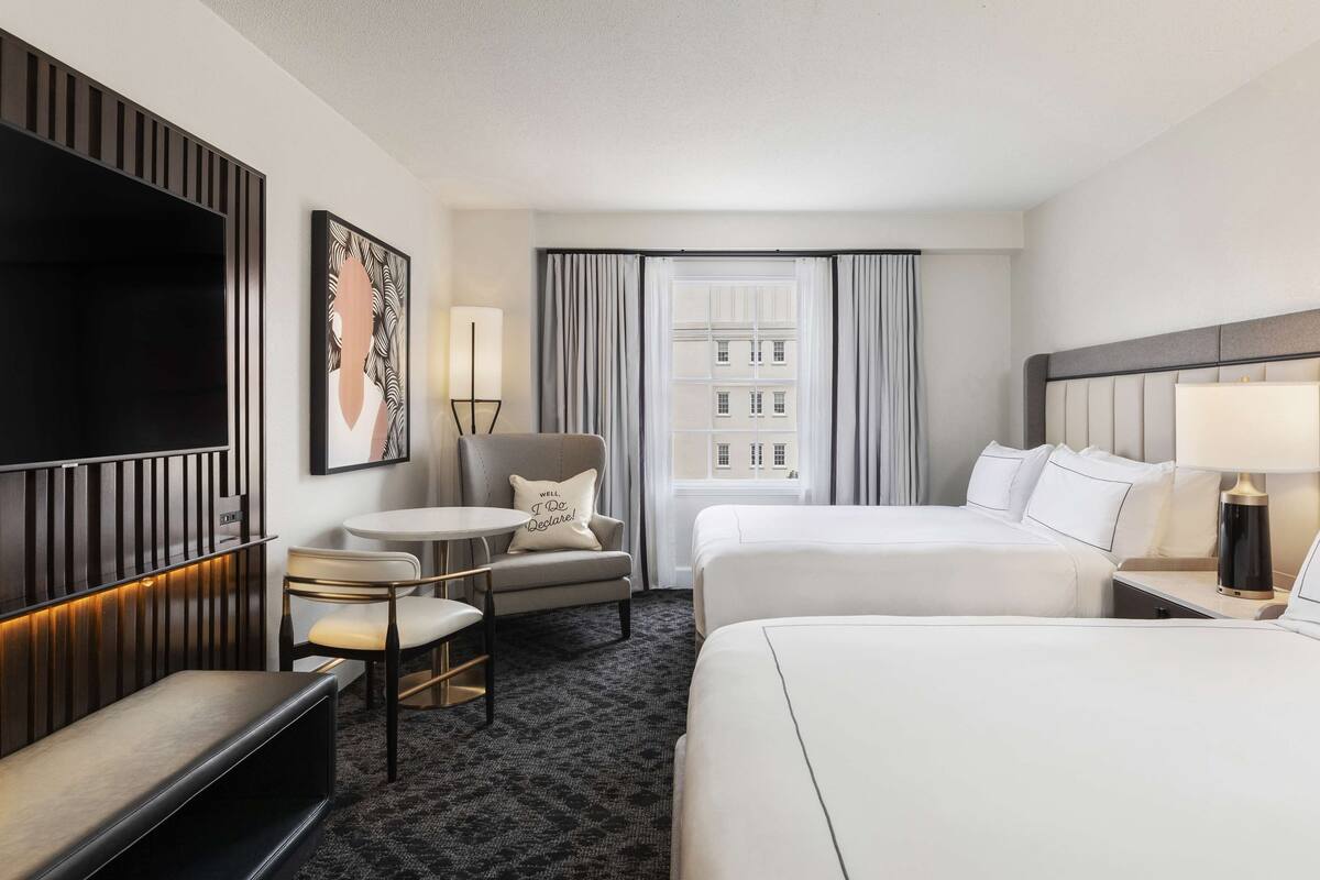 A hotel room with a large bed, a smaller bed, a desk with a lamp, a chair, a sofa, a TV, and a window with curtains.
