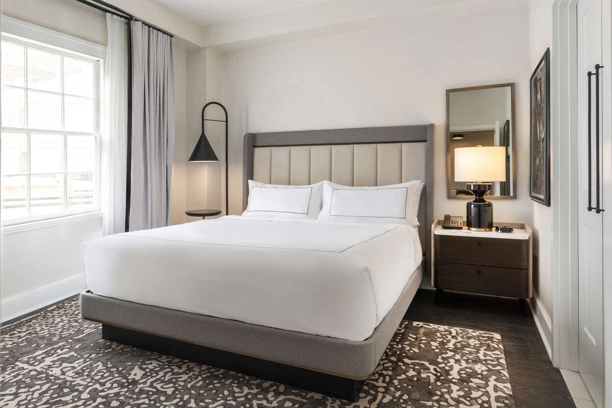 A hotel room with a large bed, a bedside table with a lamp, a mirror, and a view of the city through the window.