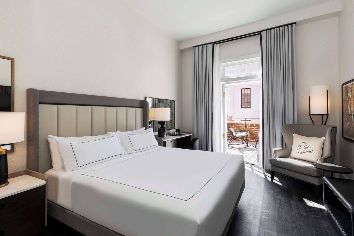 A hotel room with a large bed, a desk with a lamp, a chair with a cushion, and a view of the outside balcony.