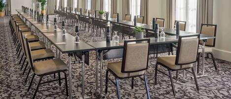 A long conference room with a patterned carpet, multiple tables, and chairs, large windows with curtains, and chandeliers.