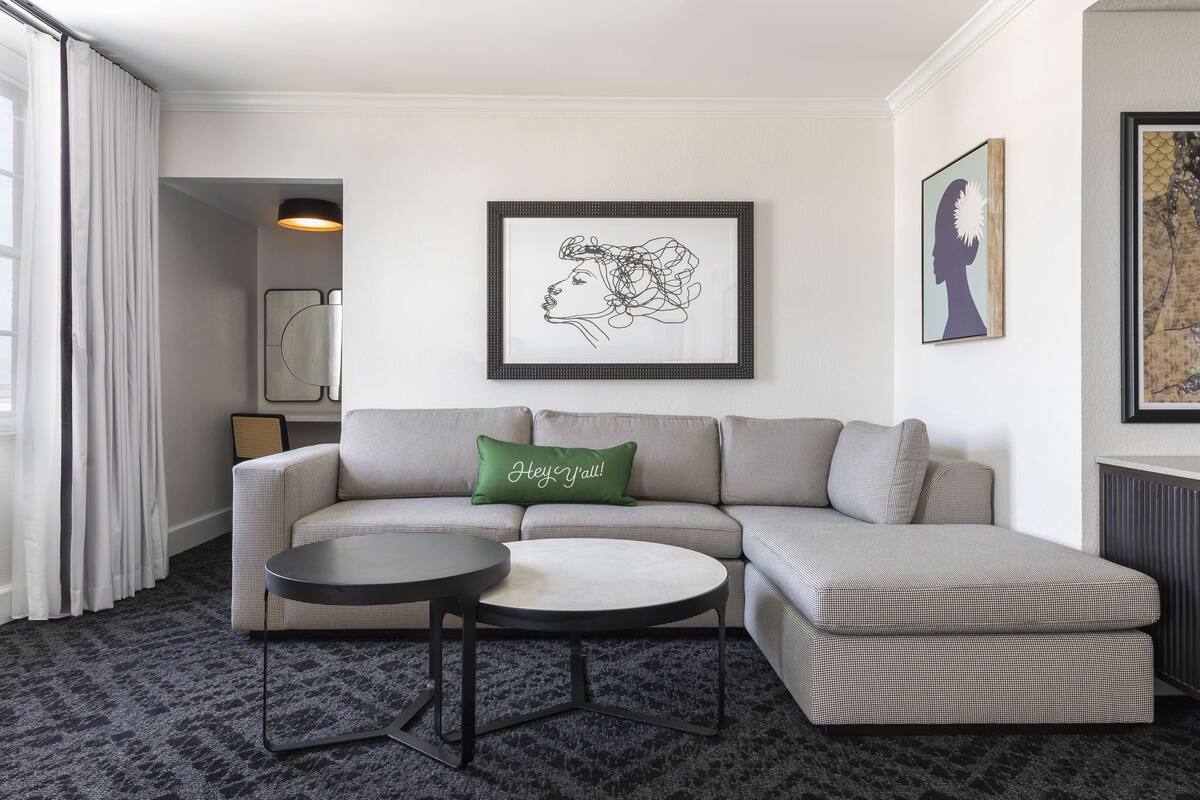 A modern living room with a checkered sofa, a round coffee table, and a green pillow with text. There are framed artworks on the walls and a radiator on the right.