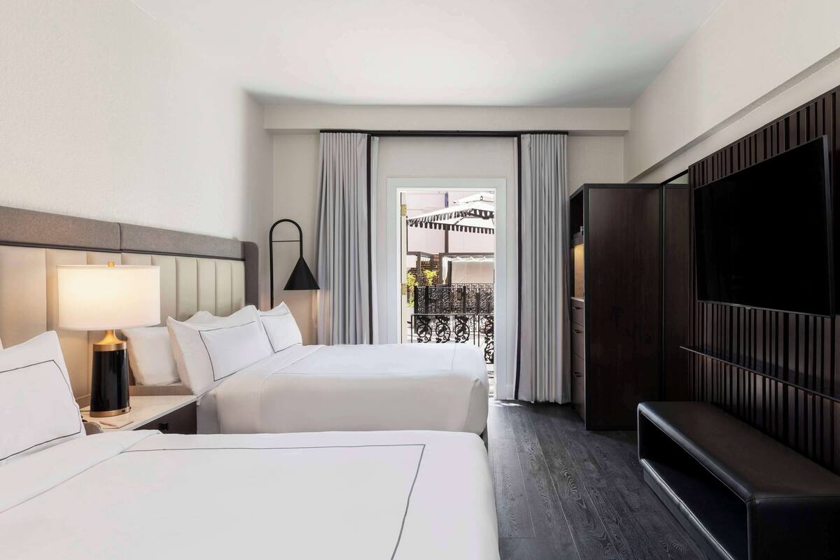 A hotel room with two beds, a TV, a desk with a lamp, and a view of an outdoor balcony.