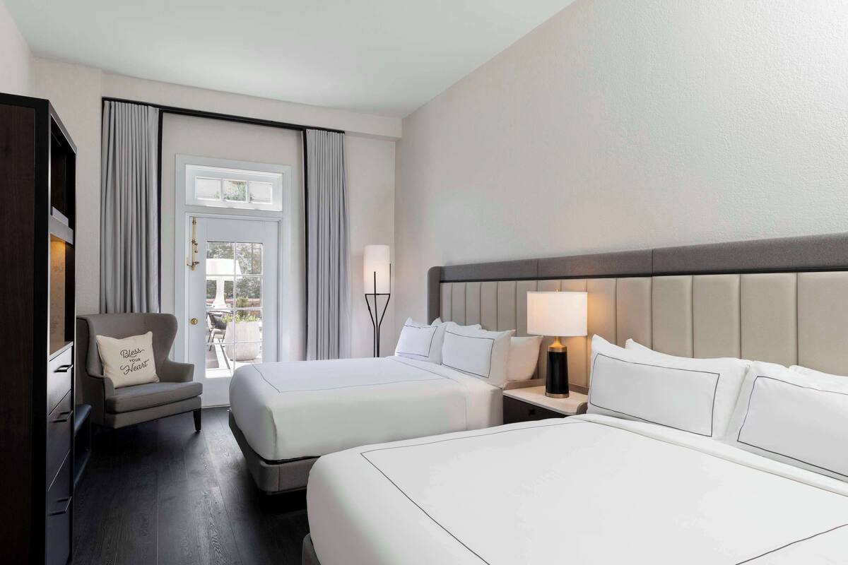 A hotel room with two beds, a chair, a nightstand with a lamp, and a view of the outdoors through a window.