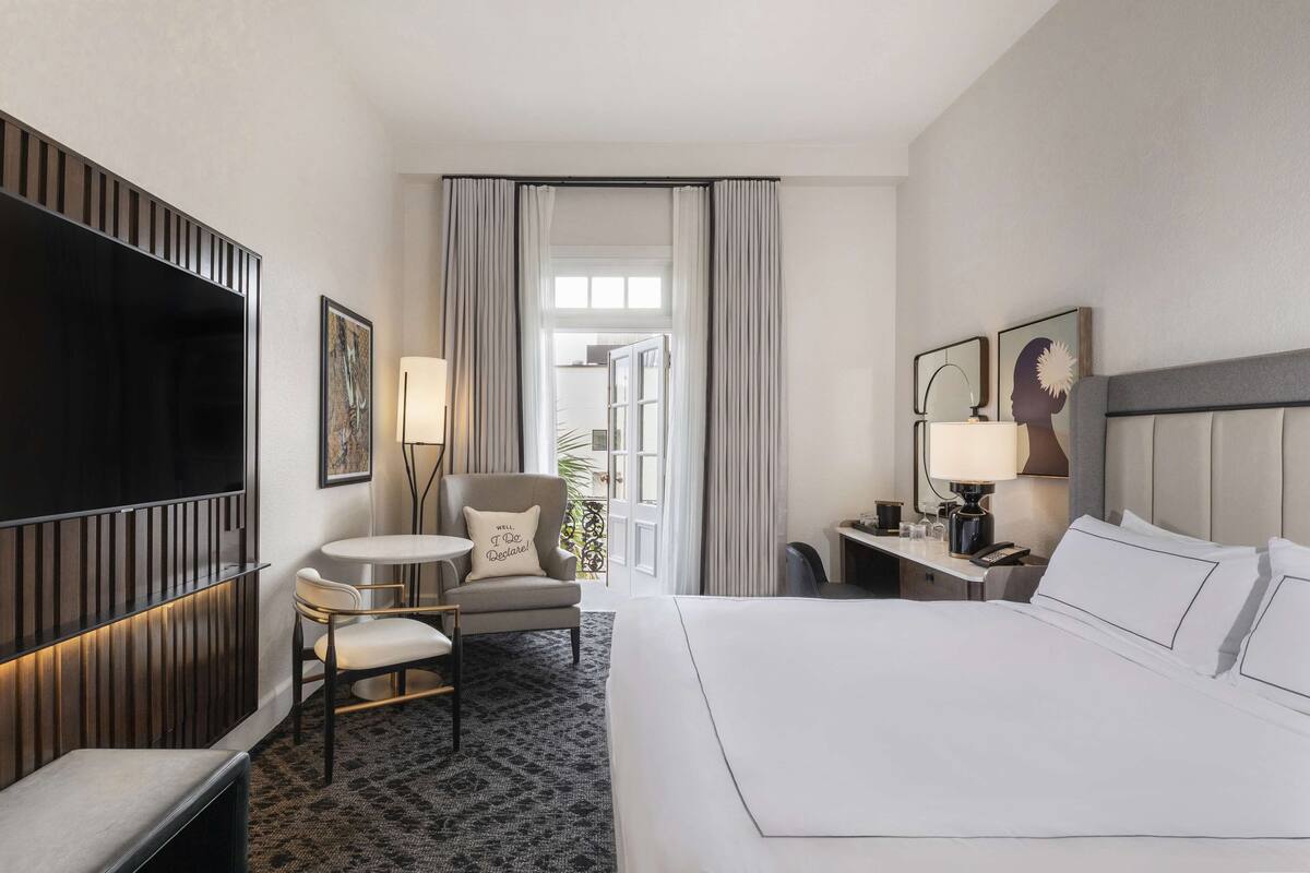 A hotel room with a large bed, a TV, a desk with a lamp, a chair, and a view of the outdoors through a window.