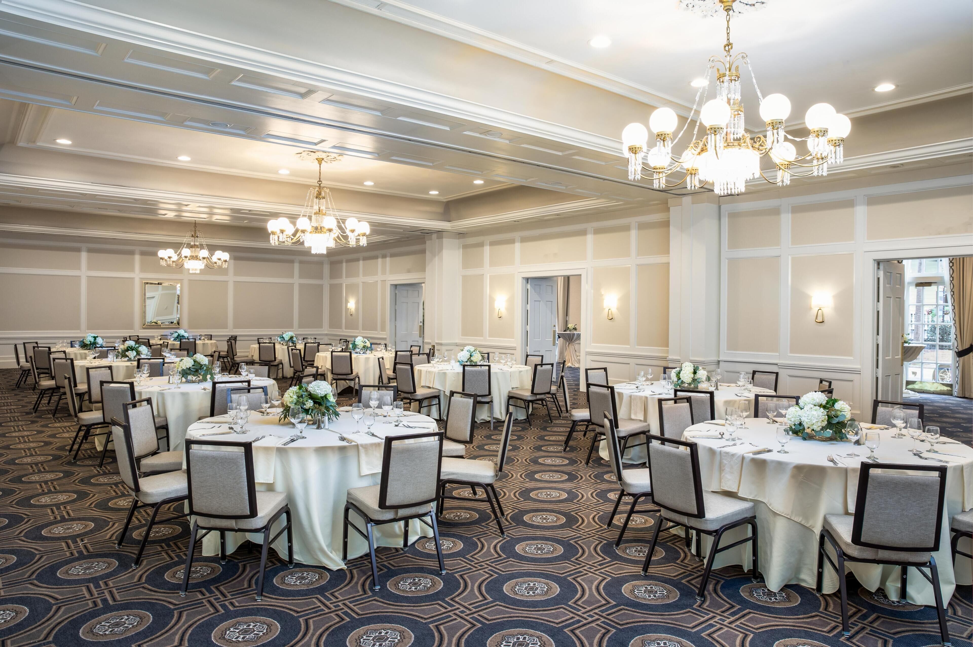 A banquet hall with round tables set for dining, chandeliers, and a patterned carpet.